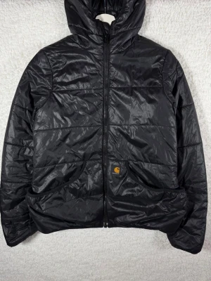 Svart pufferjacka från Carhartt - Snygg svart pufferjacka från Carhartt med glansig finish och två stora fickor framtill. Jackan har hög krage, dragkedja hela vägen och Carhartt-logga på ena fickan. Perfekt för kalla dagar och riktigt street.
