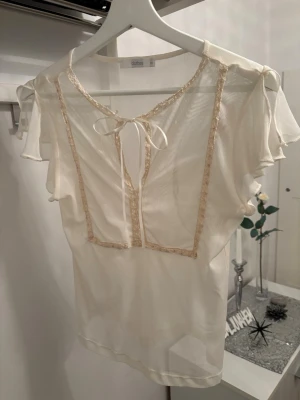 Champanjefärgad blus St.Xs - Söt champangefärgad genomskinlig blus  med volangiga korta ärmar och dekorativ spetskant framtill. Blusen har en liten rosett vid halsen och är gjord i ett tunt, lätt material som ger en romantisk känsla. Perfekt att styla med linne under.