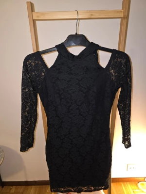 Svart spetsklänning med cut out-detaljer - Säljer en svart, figurnära spetsklänning med höghalsad design och cut out-detaljer vid axlarna. Klänningen har trekvartsärmar i genombruten spets och kort längd. Perfekt för dig som vill ha en edgy och elegant look.