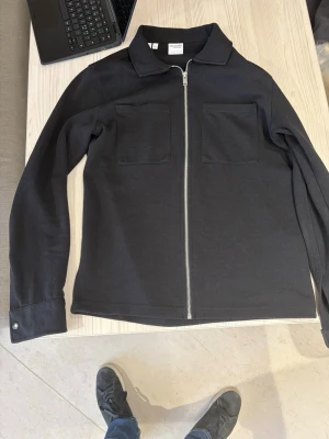 Svart overshirt selected homme - Stilren svart overshirt från Selected homme med rak passform, två stora bröstfickor och silverfärgad dragkedja framtill. Overshirten har klassisk krage och knappar vid ärmsluten. Perfekt att bära som lager över t-shirt. Materialet är mjukt och ser ut att vara bomull eller bomullsblandning.