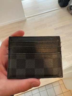 Rutigt kortfodral från Louis Vuitton - Snyggt kortfodral från Louis Vuitton i svart och grått med det klassiska rutmönstret Damier Graphite. Fodralet har flera kortfack och är gjort i tåligt läder. Perfekt för dig som vill ha en stilren och exklusiv accessoar i fickan.