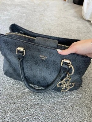 Väska GUESS - En fin och rymlig väska ifrån guess. Väskan har flera fack, bland annat tre större och några små för kort mm. Väskan har inga tydliga tecken på användning och är i väldigt fint skick