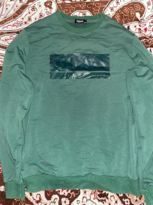 Grön sweatshirt från Hugo Boss - Snygg grön sweatshirt från Hugo Boss med stor logga i metallic på bröstet. Tröjan har rund halsringning, långa ärmar och ribbade muddar vid ärmslut och nederkant. Perfekt för en avslappnad och trendig look.