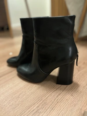 Svarta stövletter med klack - Snygga svarta stövletter i slätt läder med rundad tå och kraftig blockklack. Dragkedja baktill för enkel på- och avtagning. Perfekta för dig som vill ha en stilren och trendig look.