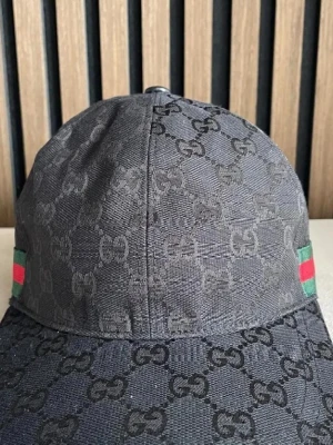 Svart Gucci keps med GG-mönster - Säljer en svart keps från Gucci med klassiskt GG-monogram över hela kepsen. På sidan finns den ikoniska gröna och röda randen. Kepsen har böjd skärm och är tillverkad i ett slitstarkt tygmaterial. Perfekt för dig som vill ha en lyxig och trendig accessoar.