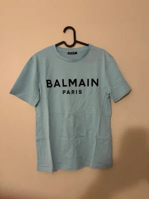 Ljusblå Balmain Paris t-shirt - Snygg ljusblå t-shirt från Balmain Paris med svart tryck framtill. Klassisk rund halsringning och korta ärmar. Tillverkad i mjuk bomull för en skön känsla och enkel stil. Direkt köpt från Dubai 