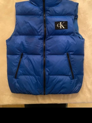 Blå dunväst från Calvin Klein  - Snygg blå dunväst från Calvin Klein Jeans med hög krage och två dragkedjeförsedda fickor framtill. Västen har ett stort CK Jeans-märke på bröstet och quiltad design som ger en puffig look. Perfekt att slänga över en hoodie för extra värme.