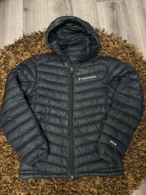 Svart dunjacka Peak Performance - Säljer en svart dunjacka från Peak Performance med huva och dragkedja framtill. Jackan har diskret logga på bröstet och är lätt vadderad med klassisk quiltad design. OBS! Dragkedja trasig