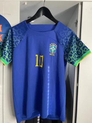 Blå Brasilien Neymar Jr matchtröja - Snygg blå Brasilien matchtröja från Nike med Neymar Jr och nummer 10 i gult på ryggen. Tröjan har coola leopardmönstrade ärmar i blått och grönt med gula detaljer. Perfekt för dig som älskar fotboll och vill sticka ut på planen eller läktaren. även ett par vita shorts med blåa ränder och nummer 10 på höger sida. Jag är 1.63 och den sitter bra på mig men rekommenderar 1,55-1,50 men funkar längre absolut,