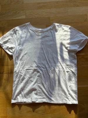 Vit basic t-shirt i bomull - En klassisk vit t-shirt i mjuk bomull med rund hals och korta ärmar. Perfekt enkel modell som passar till allt och har en avslappnad passform. Inga synliga tryck eller detaljer, bara clean och stilren.