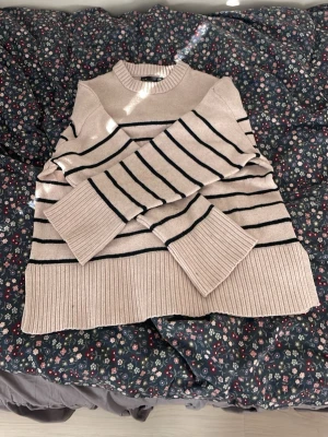 Beige stickad tröja med svartaränder - Snygg beige stickad tröja med svarta horisontella ränder och bred ribbad mudd vid ärmslut och nederkant. Tröjan har en högre rund halsringning och avslappnad passform. Perfekt för dig som gillar klassisk men trendig stil. Jätte skön til hösten. Storleken är S