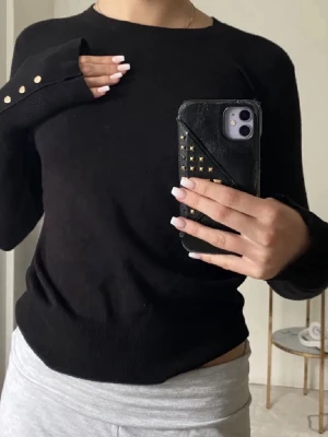 Svart stickad tröja med knappar - Svart stickad tröja med rund halsringning och långa ärmar. Ärmsluten har dekorativa guldfärgade knappar som detalj. Tröjan har en enkel och stilren design som passar till mycket. Mjuk och bekväm stickning.