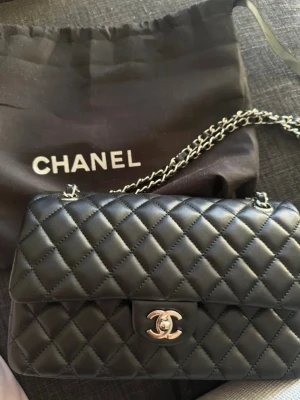 Svart Chanel quiltad handväska - Säljer en klassisk svart quiltad handväska från Chanel med silverfärgad kedja och ikonisk CC-logga på locket. Väskan är tillverkad i mjukt läder och har ett innerfack med dragkedja. Perfekt för dig som gillar tidlös och lyxig design.Säljer då den ej andvända alls, helt ny och vill bara bli av med den box o dustbag medföljer och kvitto finns på bild!