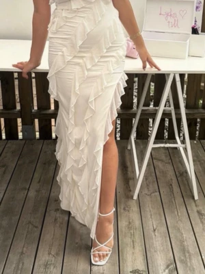 Vit långklänning med volanger och slits - Superfin vit långklänning med volangdetaljer och hög slits framtill. Klänningen är ärmlös och har en figurnära passform som framhäver siluetten. Perfekt för sommarens alla speciella tillfällen.