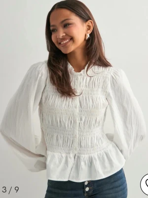 Vit blus med volang från Nelly - Supersöt vit blus från Nelly med volang nertill och smockad överdel. Ballongärmar och knappar framme. Bara använd 1 gång 