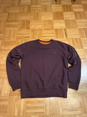 Mörkbrun alpha industries sweatshirt med rund hals - En enkel och stilren mörkbrun sweatshirt med rund hals och långa ärmar. Tröjan har ribbade muddar vid ärmslut och nederkant samt en liten diskret logga på bröstet. Insidan av kragen har en orange detalj som ger en cool kontrast.