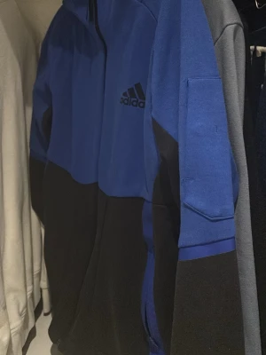 Blå och svart hoodie från Adidas - Snygg hoodie från Adidas i blått och svart med klassisk logga på bröstet. Hoodien har huva och en sportig look, samt praktisk ficka på ärmen. Tillverkad i mjukt material som känns skönt mot huden. Perfekt för dig som gillar streetwear och sportig stil.