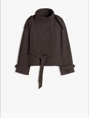 Kort brun trenchcoat med bälte - Stilren kort trenchcoat i mörkbrunt med hög krage, dubbelknäppning och axelklaffar. Jackan har ett matchande midjebälte och dekorativa knappar vid ärmsluten. Perfekt för dig som gillar klassisk men modern look.