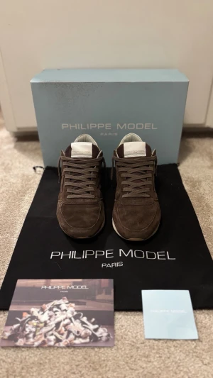Bruna Philippe Model mockasneakers - Bruna sneakers från Philippe Model i mjuk mocka med vita detaljer på häl och plös. Klassisk låg modell med snörning, vit sula och ikonisk sköldlogga på sidan. Snygg och stilren design som funkar till många olika looks.