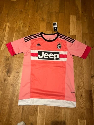 Juventus away kit 15/16 X Pogba 6 - Juventus rosa matchtröja från Adidas med Pogba och nummer 6 på ryggen. Tröjan har svarta och vita detaljer, Jeep-logga på bröstet, samt svarta ränder på axlarna. Tillverkad i lätt funktionsmaterial, perfekt för fotbollsträning.