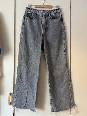 Gråa vida jeans med rå kant - Säljer ett par gråa jeans med bred passform och råa, fransiga benslut. Jeansen har klassisk femficksdesign och hög midja. Perfekta för en avslappnad och trendig look. Materialet är denim i 100% bomull.