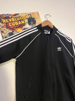 Svart Adidas ziptröja med vita ränder - Klassisk svart ziptröja från Adidas med vita ränder längs ärmarna och Adidas-logga på bröstet. Tröjan har dragkedja framtill, ribbade muddar och två fickor. Perfekt för en sportig och avslappnad stil.