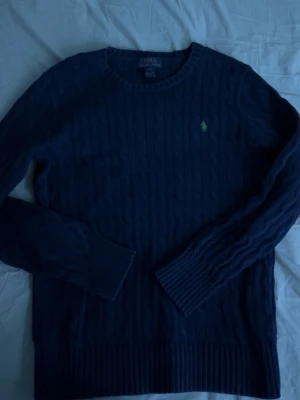 Mörkblå kabelstickad tröja Polo Ralph Lauren - Snygg mörkblå kabelstickad tröja från Polo Ralph Lauren med rund halsringning och den klassiska gröna logon broderad på bröstet. Tröjan har ribbade muddar vid ärmslut och nederkant. Perfekt för dig som gillar stilren och tidlös design.