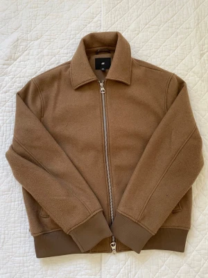 Beige ulljacka från H&M med dragkedja - Stilren beige jacka från H&M i ullblandning med klassisk krage och silverfärgad dragkedja framtill. Jackan har ribbade muddar vid ärmslut och nederkant samt två sidofickor. Perfekt för dig som gillar en clean och enkel look.