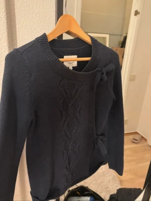 Mörkblå stickad tröja Bondelid XS - Mörkblå stickad tröja från Bondelid med dekorativa rosetter och flätstickat mönster framtill. Tröjan har rund halsringning och långa ärmar. Perfekt för dig som gillar klassisk och söt stil.