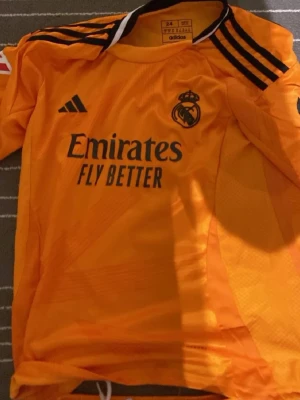 Real Madrid målvaktströja och shorts - Säljer ett orange Real Madrid målvaktsset från Adidas med matchande tröja och shorts. Tröjan har svarta detaljer, klubbmärke och sponsortryck framtill. Shortsen har nummer 5 och klubbmärke. Perfekt för fotbollsträning eller match.