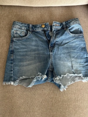 Blå jeansshorts med fransig kant - Snygga blå jeansshorts med fransig, rå kant och klassisk femficksdesign. Shortsen har normal passform och stängs med knapp och dragkedja. Perfekta för varma dagar och en avslappnad stil.