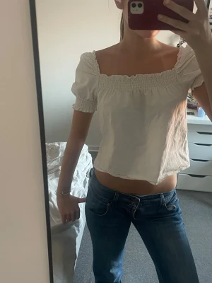 Vit croppad blus med puffärm - Söt vit blus med croppad passform och puffärmar. Toppen har smockdetaljer vid halsringning och ärmslut. Perfekt till jeans eller kjol för en trendig look. Man kan ha den offshoulder också!