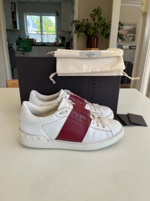 Vita valentino open sneakers med röd detalj - Snygga vita valentino open sneakers från Valentino med bred vinröd läderdetalj över snörningen och coola nitar på hälen. Skorna har en klassisk låg profil och är tillverkade i läder, vilket ger en lyxig känsla och stilren look. Skorna är i bra skick men tecken på användning och är i storlek 41. Obs att endast skor följer med! Skorna är köpa från Vestiaire collective och är självklart äkta. Vid frågor eller funderingar är det bara att säga till Mvh.