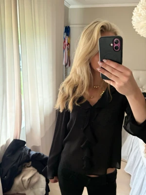 Svart blus med volangdetaljer - Snygg svart blus med v-ringning och volangdetaljer framtill. Blusen har långa ärmar och en lätt glansig finish. Blusen är från Anna Field i storlek S 