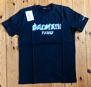Svart Balmain Paris t-shirt - Snygg svart t-shirt från Balmain med ett stort vitt Balmain Paris-tryck i en cool, lite sliten stil på bröstet. Klassisk rund halsringning och korta ärmar. Perfekt för dig som vill ha en statement-tee från ett exklusivt märke.