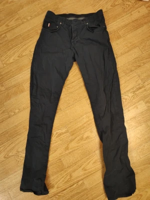 Mörkgrå jeans byxor med rak passform stl 30/34 - Snygga mörkgrå jeans med klassisk femficksdesign och rak passform. Byxorna har en liten detalj med röd, vit och blå rand på myntfickan. Perfekta för dig som gillar en stilren och enkel look. Stl herr 30/34