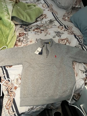 Grå halvzip från Polo Ralph Lauren - Snygg grå sweatshirt med halv dragkedja från Polo Ralph Lauren. Tröjan har en klassisk passform, ribbade muddar och den ikoniska röda loggan broderad på bröstet. Perfekt för lager-på-lager och chill stil.