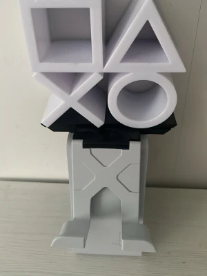PlayStation handkontrollställ vit - Snyggt handkontrollställ från PlayStation i vit plast med de klassiska symbolerna fyrkant, triangel, cirkel och kryss. Perfekt för att hålla din handkontroll på plats och ge gaminghörnan en cool look.