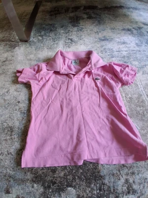 Rosa pikétröja från Lacoste - Snygg rosa pikétröja från Lacoste med klassisk krage och korta ärmar. Tröjan har den ikoniska krokodilloggan på bröstet och är tillverkad i mjuk bomull. Perfekt för en avslappnad och sportig stil.