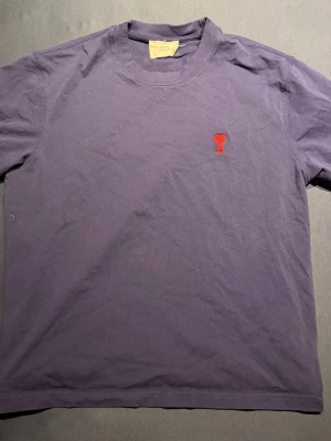 Blå t-shirt från Ami Paris - Snygg blå t-shirt från Ami Paris med röd broderad logga på bröstet. Klassisk passform och rund halsringning. Tillverkad i mjuk bomull som känns skön mot huden. Perfekt för en clean och stilren look.