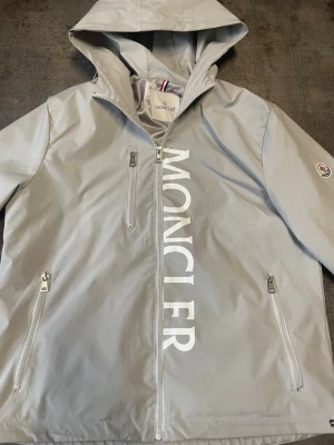 Ljusgrå vindjacka från Moncler - Snygg ljusgrå vindjacka från Moncler med stor vit logga längs dragkedjan. Jackan har huva, tre dragkedjefickor och Moncler-märke på ärmen. Tillverkad i ett lätt och vattenavvisande material, perfekt för blåsiga dagar.