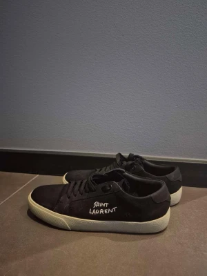 Svarta Saint Laurent sneakers - Saint Laurent sneakers i svart – stilren designerlook Snygga och minimalistiska Saint Laurent-sneakers i svart med klassisk low-top design som passar till allt. Bekväma och enkla att matcha, både till jeans och mer uppklädda outfits. Skorna är använda och har visst slitage enligt bilderna, men har fortfarande mycket stil kvar. Säljes då de inte används längre. Kolla gärna alla bilder för skick och detaljer. Påsen till skorna följer såklart också med.  