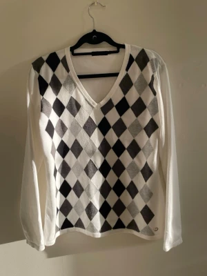 Vit argyle stickad tröja - Vit stickad tröja från Röhnisch med klassiskt argylemönster i svart och grått framtill. Tröjan har V-ringning och långa ärmar. Materialet är tunt och mjukt, perfekt för lager-på-lager. Diskret logga nertill på framsidan.