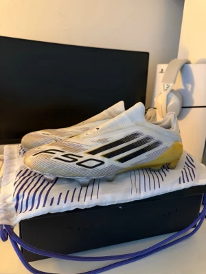 Adidas F50 Elite LL FG fotbollsskor - Säljer ett par Adidas F50 Elite LL FG fotbollsskor i vitt med svarta och guldiga detaljer. Skorna har en strömlinjeformad design utan snörning och är tillverkade i syntetmaterial för bästa prestanda på gräsplan. Perfekta för dig som vill sticka ut på planen.