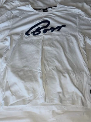 Vit t-shirt från BOSS med blå logga - Snygg vit t-shirt från BOSS med stor blå broderad logga på bröstet. Klassisk rund hals och korta ärmar. Tillverkad i mjuk bomull som känns skön mot huden. Perfekt för en clean och stilren look.