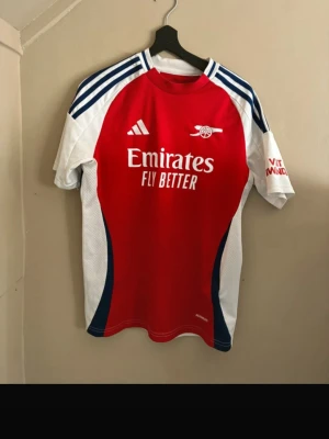 Arsenal matchtröja Adidas röd/vit - Snygg Arsenal matchtröja från Adidas i rött och vitt med blå detaljer. Tröjan har klubbmärke och Emirates Fly Better-tryck på bröstet samt Vitamina Well-logga på ärmen. Tillverkad i lätt och ventilerande material, perfekt för fotboll eller träning.