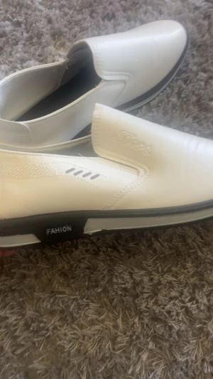 Vita loafers med svart sula - Säljer ett par vita loafers med svart sula och diskreta detaljer på sidan. Skorna har elastiska paneler för enkel påtagning och texten 'FASHION' på sulan. Materialet ser ut att vara syntetiskt läder och designen är stilren och modern. dom kostade över 1000 och jag bara används en gång