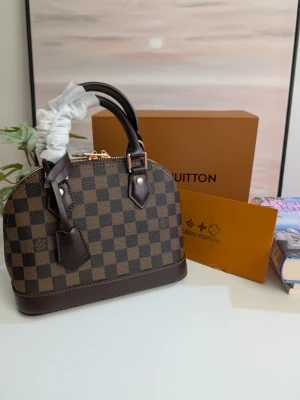 Iouis Vuitton Alma BB 🤎  - Säljer denna otroligt fina och eleganta handväska i en tidlös "dome/skalmodell". Väskan är helt oanvänd och i absolut nyskick – skyddsplasten sitter till och med kvar runt handtaget! Detaljer: Mönster: Snyggt schackrutigt mönster i mörkbruna och ljusbruna toner. Detaljer: Eleganta detaljer i guldmetall och mörkbrunt material på botten och handtag. Tillbehör: Kommer med en matchande namntagg framtill samt tillhörande låda och skyddstyg (se bild). Skick: 10/10 – Helt ny och aldrig använd. 