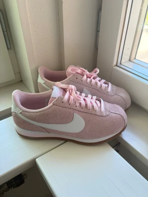 Nike Cortez rosa sneakers - Säljer ett par Nike Cortez sneakers i ljusrosa mocka med vita detaljer och klassisk vit swoosh på sidan. Skorna har rosa snören, vit mellansula och yttersula i gummi. Perfekta för dig som vill ha en retro och stilren look. Modellen är sällsynt och skorna är enbart använd fåtalet gånger.