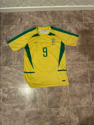 Brasilien Ronaldo 9 matchtröja Nike - Gul Brasilien fotbollströja från Nike med gröna detaljer och nummer 9 på bröstet och ryggen. RONALDO står tryckt på ryggen. Klassisk design med korta ärmar och rund hals, tillverkad i lätt och ventilerande material.